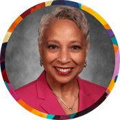 Portrait of a woman with medium-brown skin and short gray hair, smiling in a pink blazer. Set against a gray background with a colorful circular border.