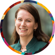 Smiling woman with pale skin and straight chestnut-brown hair, wearing a green top. Set against a soft outdoor background, framed in a multicolor circle.