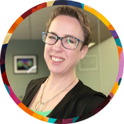 A smiling fair-skinned woman with short light-brown hair, wearing rectangular glasses and a delicate multi‑chain necklace over a black top. They’re photographed from the shoulders up against a softly blurred indoor background featuring a framed picture on the wall. The circular frame of the image is edged with a colorful, rainbow-inspired border.