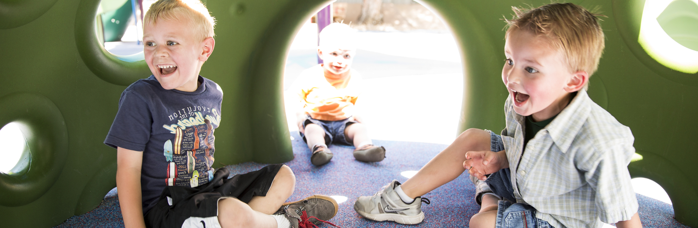 Three young children are sitting are sitting under a play structure with a look of excitement on their faces